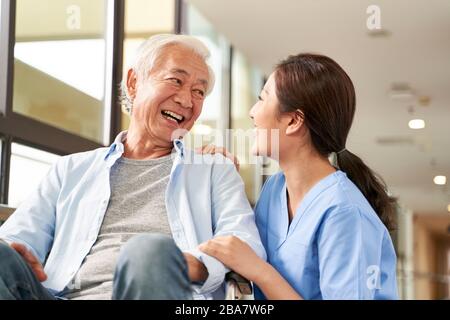 young friendly asian female caregiver talking chatting to happy senior man in hallway of nursing home Stock Photo