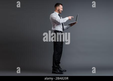 profile of angry businessman in suit pointing with finger at laptop on grey Stock Photo