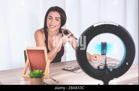 Young influencer woman creating social media videos with smartphone while putting on make up - New digital jobs trends, technology, cosmetics and fash Stock Photo