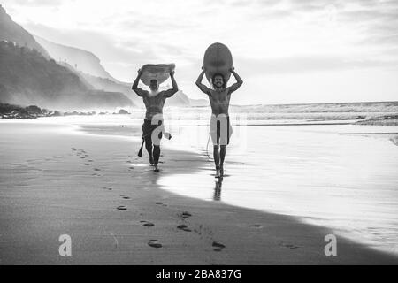 Multigeneration friends going to surf on tropical beach - Family people having fun doing extreme sport - Main focus on young man face - Black and whit Stock Photo