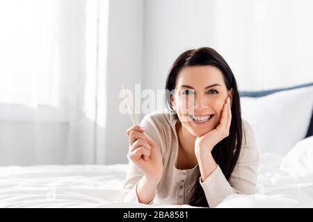 cheerful and pregnant woman holding pregnancy test and lying on bed Stock Photo