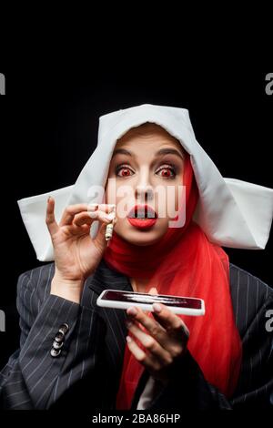 shocked demonic nun holding dollar banknote and smartphone with cocaine lines isolated on black Stock Photo