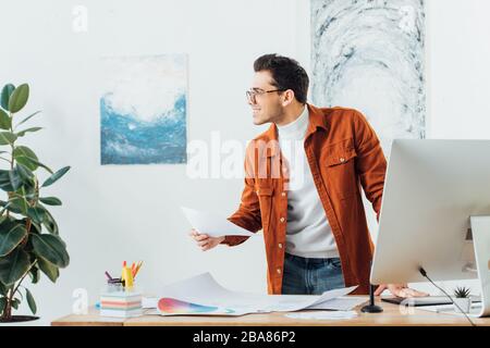 Smiling designer looking away while working on project of ux design near color circles and sketches on table Stock Photo