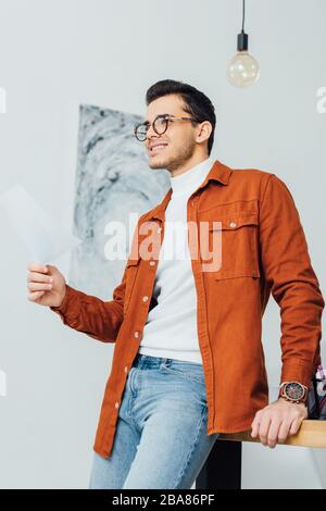 Low angle view of smiling ux designer holding paper and looking away near working table Stock Photo