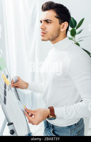 Side view of pensive ux developer holding app layouts near whiteboard in office Stock Photo