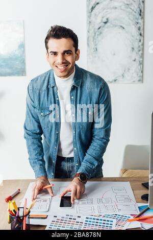 Smiling designer looking at camera while working with ux wireframe sketches, color palettes and smartphone on table Stock Photo
