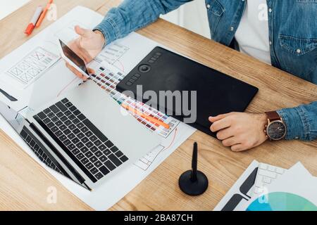 Cropped view of ux designer using smartphone near laptop, graphics tablet and sketches on table Stock Photo