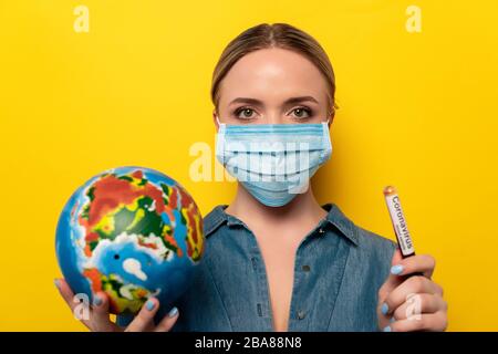 young woman in medical mask holding test tube with coronavirus blood sample and globe on yellow background Stock Photo