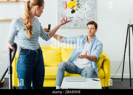 Selective focus of woman with vacuum cleaner quarreling with boyfriend holding remote controller on couch Stock Photo