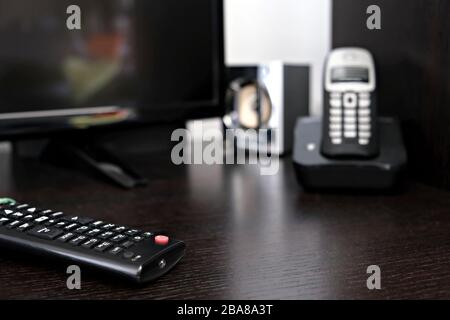 technology composition: TV remote controller in the foreground (in focus) and speaker, cordless phone an TV on the blurred background Stock Photo