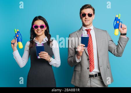 two happy businesspeople in sunglasses holding water guns, passports and air tickets on blue background Stock Photo