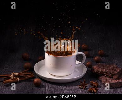 A cup with coffee splash with macadamia nuts, chocolate and cinnamon stick on table wood over dark background Stock Photo
