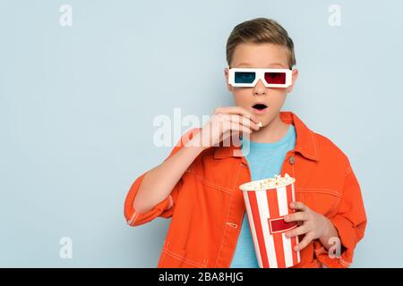 shocked kid with 3d glasses eating popcorn on blue background Stock Photo