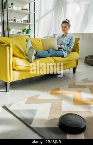 attractive woman using laptop on sofa while robotic vacuum cleaner washing carpet in living room Stock Photo