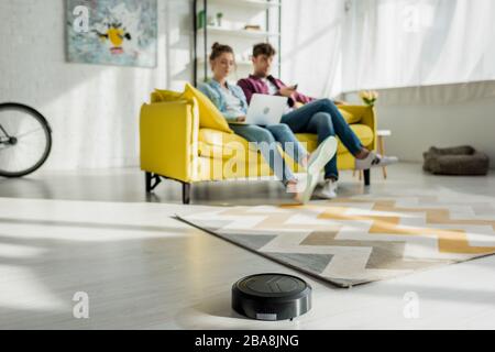 selective focus of robotic vacuum cleaner washing carpet near man watching movie and woman using laptop in living room Stock Photo