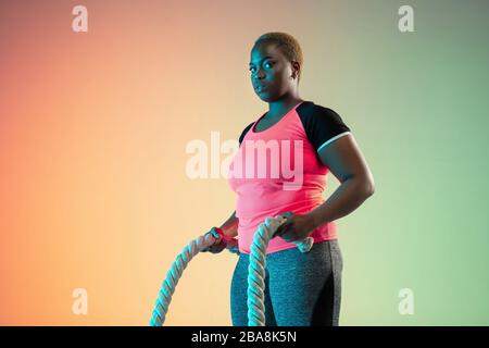 Young african-american plus size female model's training on gradient background in neon light. Doing workout exercises with the ropes. Concept of sport, healthy lifestyle, body positive, equality. Stock Photo
