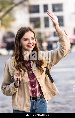 Beautiful woman waving hand and smiling with backpack in city Stock Photo