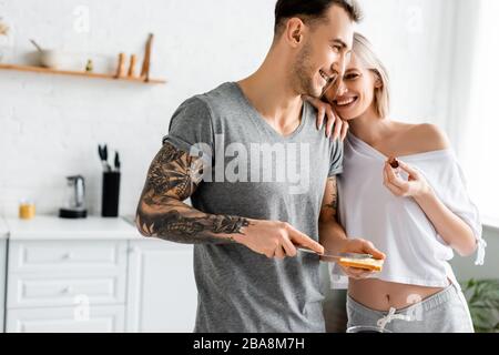 Beautiful smiling woman holding grape and embracing tattooed boyfriend with toast in kitchen Stock Photo