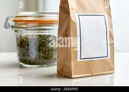 Brown kraft paper bag with white blank label in front of a bale glass jar and orange rubber gasket with dried herbs or tea on a white background. Stock Photo