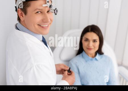 selective focus of smiling otolaryngologist in ent headlight holding ear speculum near smiling woman sitting in medical chair Stock Photo