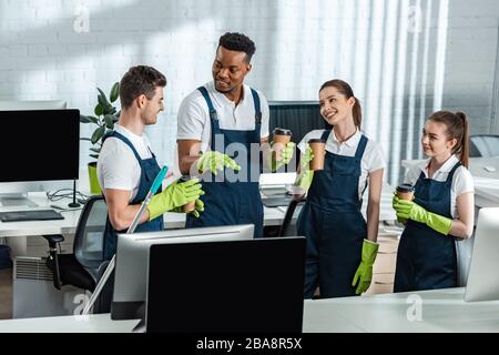 happy multicultural cleaners talking while drinking coffee to go in office Stock Photo