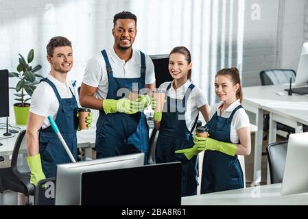 cheerful multicultural cleaners looking at camera while holding coffee to go in office Stock Photo