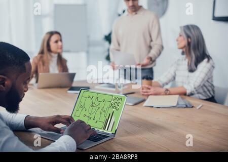 selective focus of african american man using laptop with graphs and his colleagues talking on background Stock Photo