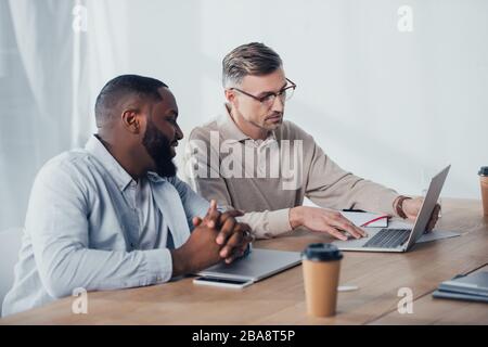 businessman using laptop and talking with smiling african american colleague Stock Photo