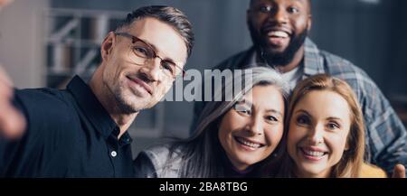 panoramic shot of smiling multicultural colleagues looking at camera and taking selfie Stock Photo