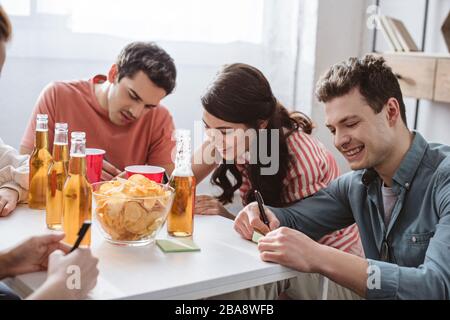 cheerful friends writing on sticky notes while playing name game at table with drinks and chips Stock Photo