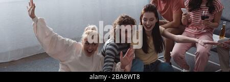 panoramic shot of cheerful girl waving hand while sitting with friends at home Stock Photo