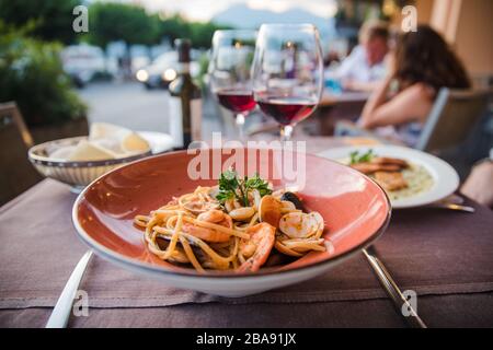 Delicious italian seafood pasta in red plate with two glasses of red wine in outdoor dinner restaurant in Bellagion town in Italy Stock Photo