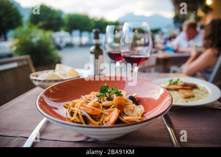 Delicious italian seafood pasta in red plate with two glasses of red wine in outdoor dinner restaurant in Bellagion town in Italy Stock Photo