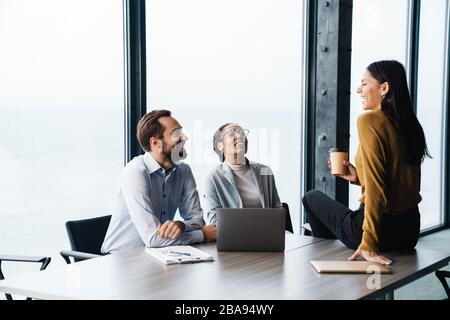 Image of multiethnic young female and male colleagues sitting by table and working on laptops in office Stock Photo
