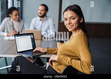 Image of multiethnic young female and male colleagues sitting by table and working on laptops in office Stock Photo