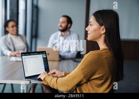 Image of multiethnic young female and male colleagues sitting by table and working on laptops in office Stock Photo