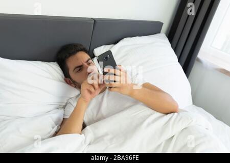 A young man in pajamas uses a mobile phone while lying in bed. Dependence on social networks and virtual communication. Stock Photo