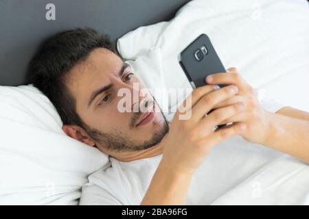A young man in pajamas uses a mobile phone while lying in bed. Dependence on social networks and virtual communication. Stock Photo