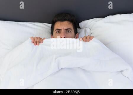 A young man is hiding in bed under a blanket at home. Fear and surprise are visible in his eyes. Stock Photo
