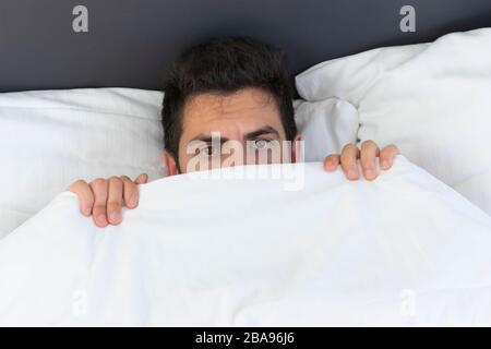A young man is hiding in bed under a blanket at home. Fear and surprise are visible in his eyes. Stock Photo
