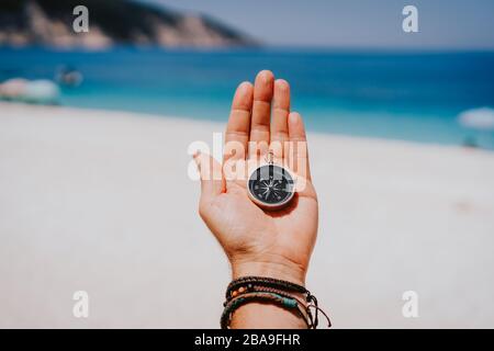 Stretched hand palm with black metal compass against summer beach and blue sea. Follow your way, goal, wish concept. Point of view pov. Stock Photo