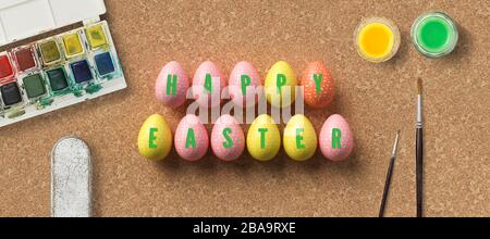 easter eggs with message HAPPY EASTER surrounded by brushes and water color boxes on cork background Stock Photo