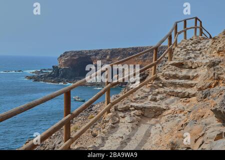 Views from the wooden staircase of a lookout with a boat sailing in the sea and the Cuevas de Ajuy in the background in Fuerteventura, Canary Islands, Stock Photo