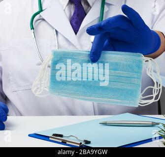 doctor in a white coat and blue latex gloves sits at a white desk and shows a stack of disposable medical masks, protective agent against viruses and Stock Photo