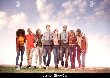 Group of diverse young people walking arm around shoulders. Millennial generation teamwork having fun bonding talking together against a cloudy sky. C Stock Photo