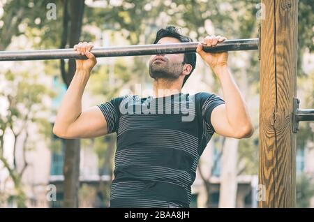 Young man making pull-up strength training exercise - fitness guy working out his arm muscles on outdoor park gym doing chin-ups / pull-ups CrossFit Stock Photo