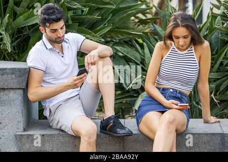 End of a relationship. A couple breaking up their love story, sitting on a park in the summer and watching their smartphones. Relationship and technol Stock Photo