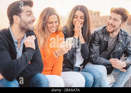 Group of four friends having a fun together. Two women and two men sitting on a bench talking laughing and enjoying their time using smartphone. Stock Photo
