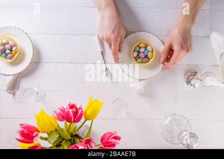 Top view male hands serving Easter table with colorful sweet eggs in cases on the plates. Crystal glassware and fresh tulip flowers on the white woode Stock Photo