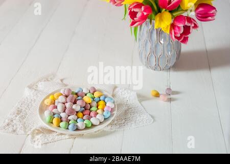 Festive Easter candies covered eggs in various pastel colors on the plate with vintage napkin near vase with fresh tulips on wooden table. Family holi Stock Photo